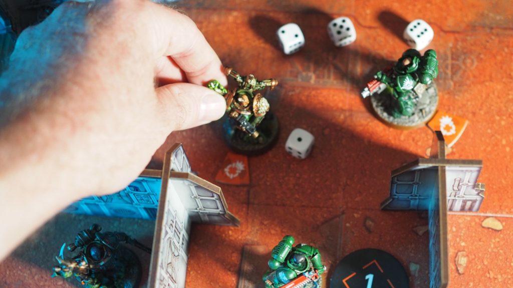 A hand places a model onto an orange battlemat with tokens, terrain, and dice