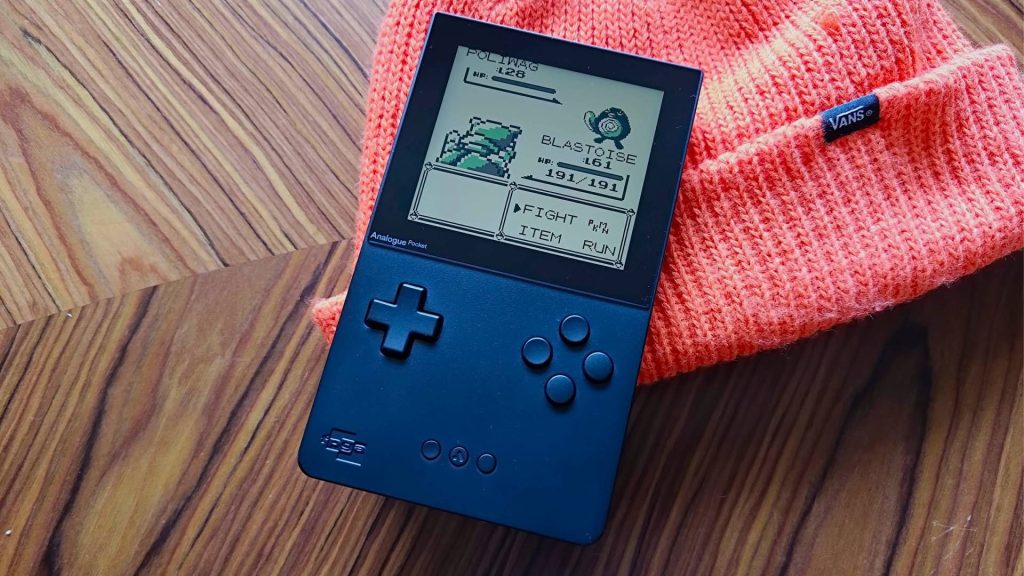 Analogue Pocket resting against orange beanie hat on woodgrain table with Pokemon Blue battle on screen featuring Blastoise and Polywag.